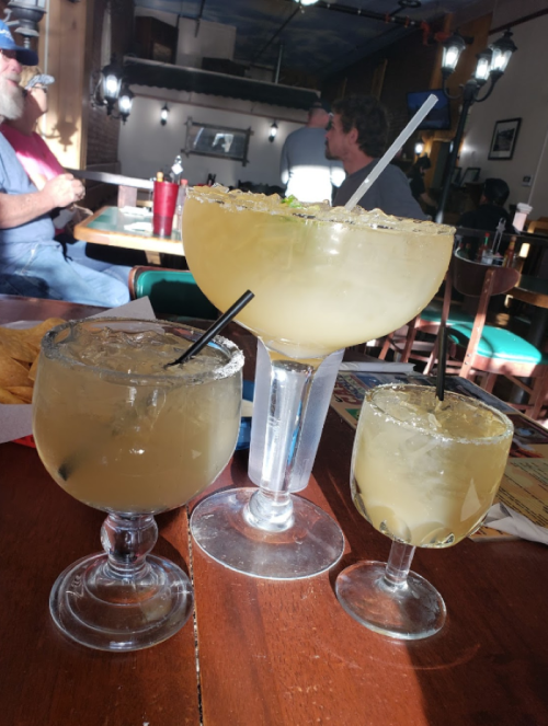 Three margaritas of varying sizes on a wooden table, with a casual dining atmosphere in the background.
