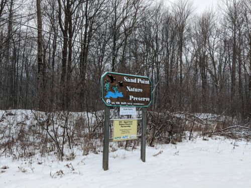 Sign for Sand Point Nature Preserve, surrounded by bare trees and snow, indicating the preserve's location and rules.