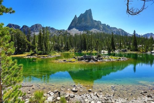 A serene lake surrounded by lush trees, reflecting a towering mountain under a clear blue sky.
