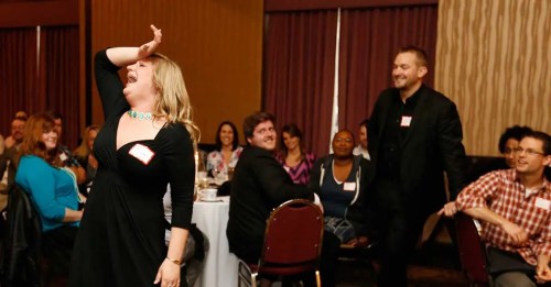 A woman in a black dress laughs joyfully while a man in a suit stands nearby, with a lively audience in the background.