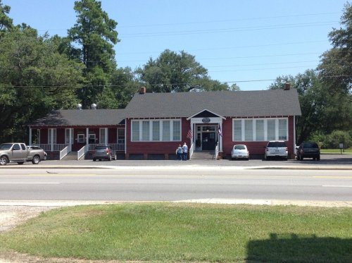 A red building with large windows, surrounded by trees, and parked cars in front on a sunny day.