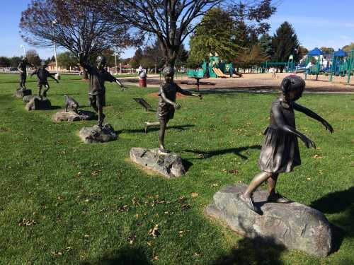 A row of bronze child statues playfully running on rocks in a park, with a playground visible in the background.