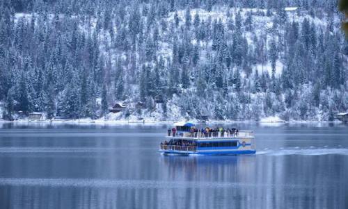 A boat with passengers glides across a snowy lake, surrounded by evergreen trees and a winter landscape.