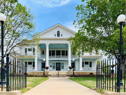 A large, historic mansion with white columns, surrounded by trees and a black iron fence, under a blue sky.