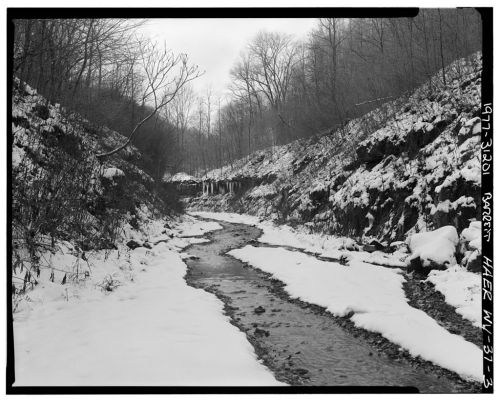 A snowy landscape with a winding creek surrounded by bare trees and snow-covered banks. Black and white photograph.
