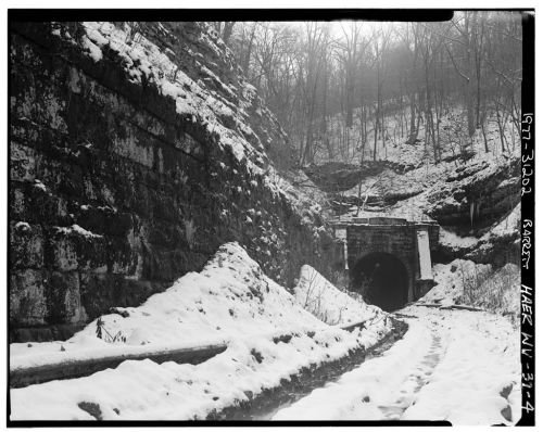 A snowy landscape featuring a stone tunnel entrance surrounded by rocky terrain and trees in the background.