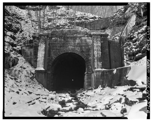 A snow-covered tunnel entrance surrounded by rocky terrain and trees, captured in black and white.