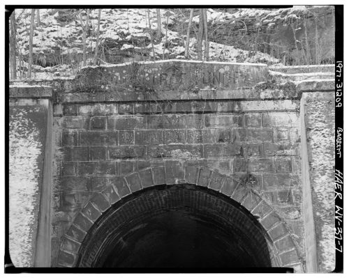 Black and white photo of a stone tunnel entrance with weathered lettering above, surrounded by rocky terrain.