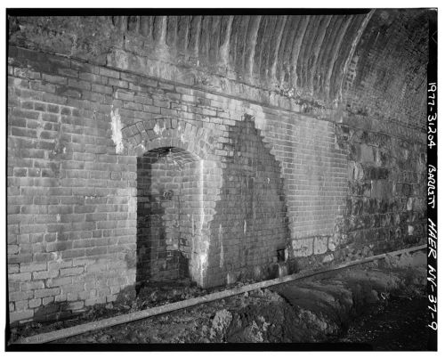 Black and white photo of a brick wall with an arched doorway and uneven surface, showing signs of wear and age.