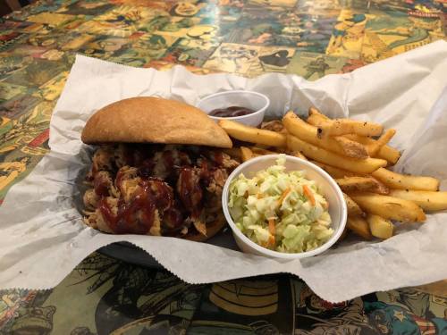 A plate with a pulled pork sandwich, coleslaw, barbecue sauce, and a side of golden fries on a colorful comic-themed table.