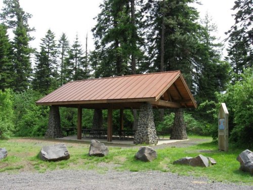 A wooden pavilion with a metal roof, supported by stone pillars, surrounded by trees and large rocks.