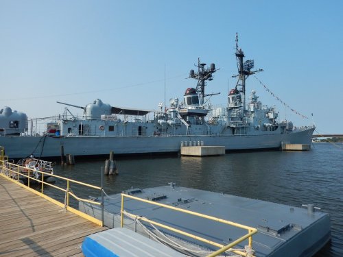 A gray naval ship docked at a pier, with flags flying and calm water surrounding it.