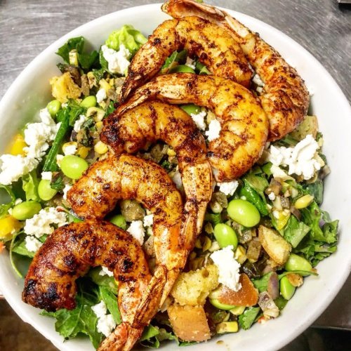 A vibrant salad topped with grilled shrimp, mixed greens, corn, edamame, and crumbled cheese in a white bowl.