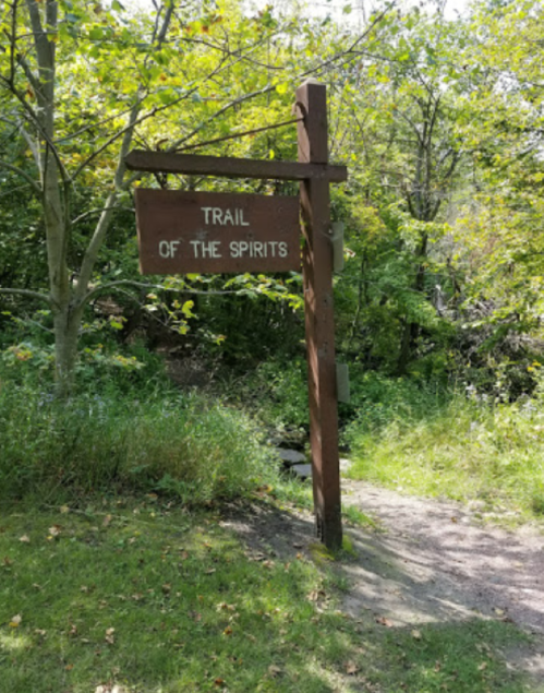 A wooden sign reading "Trail of the Spirits" stands at the entrance to a lush, green forest path.