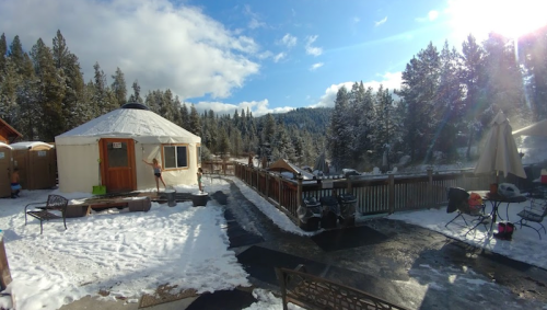 A snowy landscape featuring a yurt, surrounded by trees and a deck with outdoor furniture under a bright sky.
