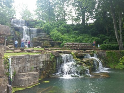 A scenic waterfall surrounded by lush greenery, with people enjoying the view on a sunny day.