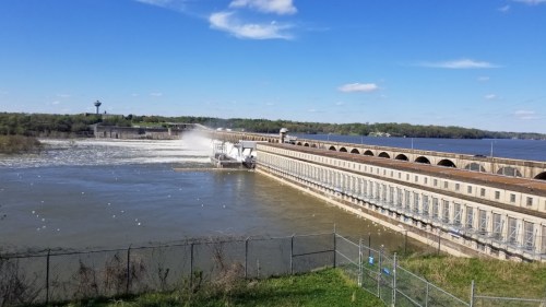A large dam with water flowing over it, surrounded by greenery and a clear blue sky with scattered clouds.