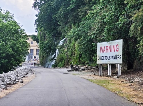 A road lined with trees and rocks, featuring a large warning sign about dangerous waters ahead.
