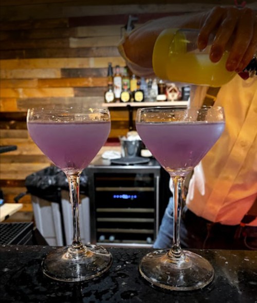 Two elegant cocktails in stemmed glasses, one being poured from a shaker, set against a rustic wooden bar backdrop.