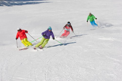 Four skiers in colorful outfits navigate a snowy slope, creating trails in the fresh powder.