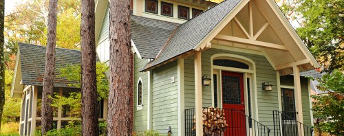A charming green house with a red door, surrounded by tall trees and a stack of firewood on the porch.