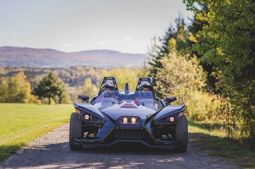 A sleek black sports car parked on a gravel path, surrounded by trees and mountains in the background.