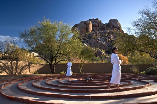 Two individuals in white robes walk through a circular stone labyrinth, with a rocky landscape in the background.