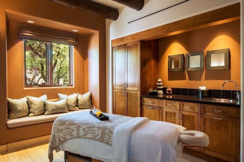 A serene spa room featuring a massage table, wooden cabinetry, and a cozy window seat with decorative pillows.