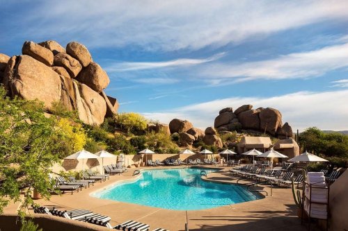 A serene pool area surrounded by large boulders and lounge chairs under umbrellas, with a clear blue sky above.