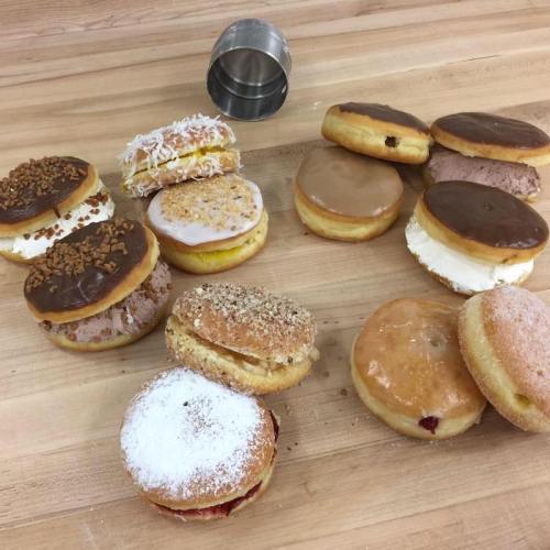 A variety of donuts on a wooden surface, including frosted, filled, and topped with coconut and nuts.