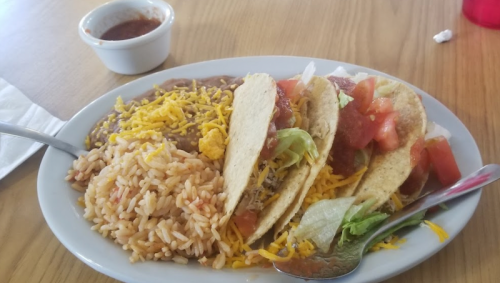 A plate of tacos with lettuce, tomato, and cheese, served with rice, refried beans, and a small bowl of salsa.