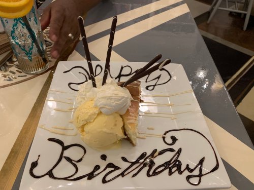 A birthday dessert plate with cheesecake, ice cream, whipped cream, and chocolate sticks, decorated with "Happy Birthday" in chocolate.