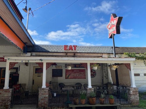 A rustic diner with a red "EAT" sign, outdoor seating, and a vintage Coca-Cola advertisement under a blue sky.
