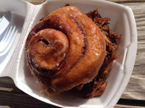 A cinnamon roll topped with icing, resting on a bed of pulled pork in a takeout container.