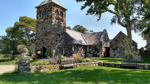 A stone church with a bell tower surrounded by greenery and benches on a sunny day.