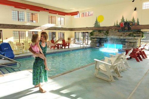 A woman holds a child by an indoor pool with a waterfall feature and colorful chairs around the area.