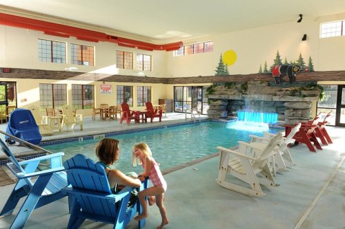 A bright indoor pool area with colorful chairs, a waterfall feature, and a child interacting with an adult.