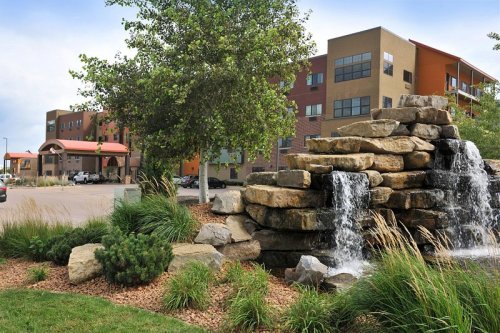 A stone waterfall surrounded by greenery, with modern buildings in the background under a cloudy sky.