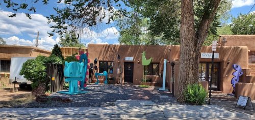 Colorful sculptures in front of a brown adobe building under a blue sky with scattered clouds.
