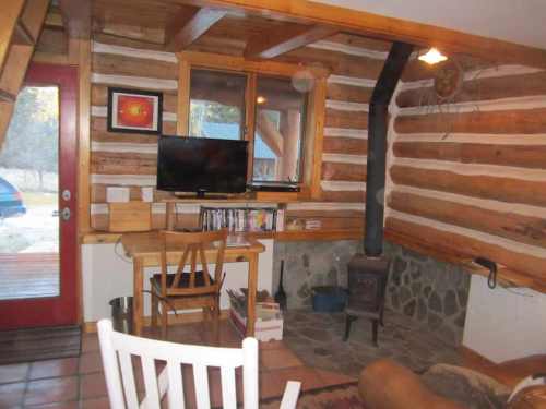 Cozy log cabin interior with a wooden table, TV, and a small stove, featuring rustic decor and a red door.