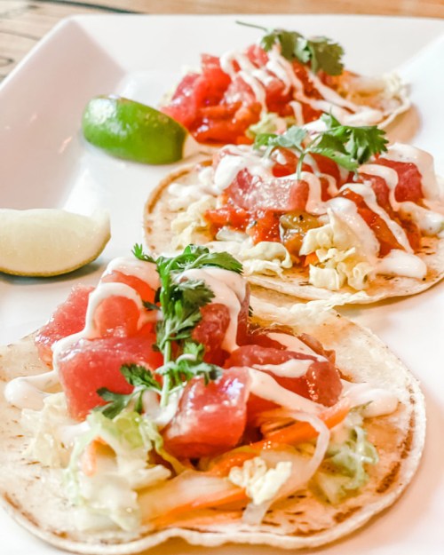 Three tacos topped with diced tomatoes, lettuce, and drizzled with sauce, served with lime wedges on a white plate.