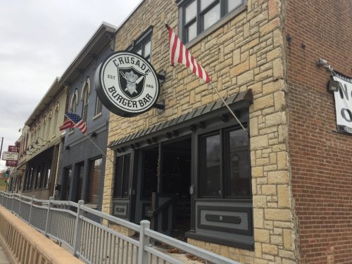 Exterior of Crusade Burger Bar, featuring a stone facade and American flags, on a cloudy day.