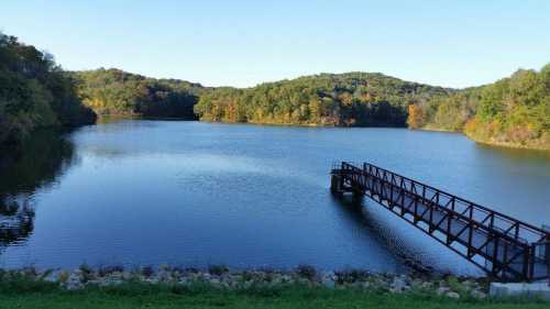 A serene lake surrounded by hills, with a wooden dock extending into the calm water under a clear blue sky.