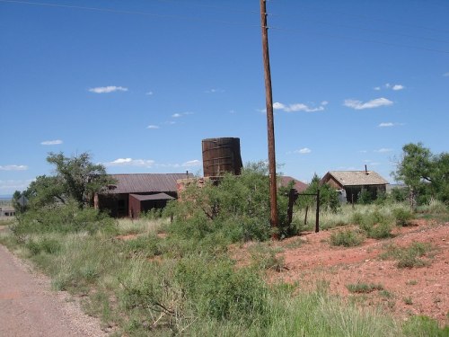 Abandoned buildings and a water tower surrounded by overgrown grass under a clear blue sky.