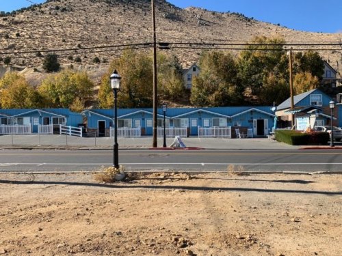 A row of blue buildings with white fences along a road, set against a hilly landscape with sparse vegetation.