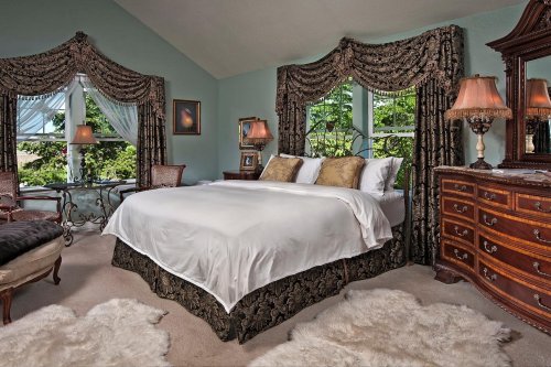 Elegant bedroom featuring a large bed, decorative curtains, vintage furniture, and soft rugs. Natural light from windows.