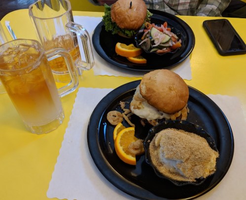 Two plates with burgers, sides of salad, and orange slices, alongside a glass of iced tea on a yellow table.