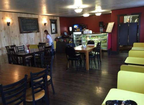 Cozy restaurant interior with wooden tables, chairs, and a counter displaying food, featuring warm lighting and a welcoming atmosphere.