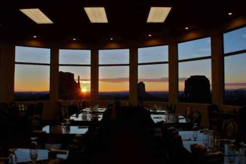 A restaurant with tables set for dining, featuring large windows showcasing a sunset over a desert landscape.