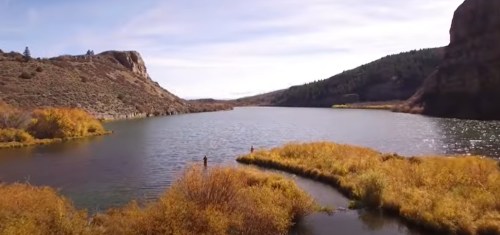 A serene lake surrounded by golden grasses and rocky hills under a clear blue sky. Two figures stand by the water's edge.